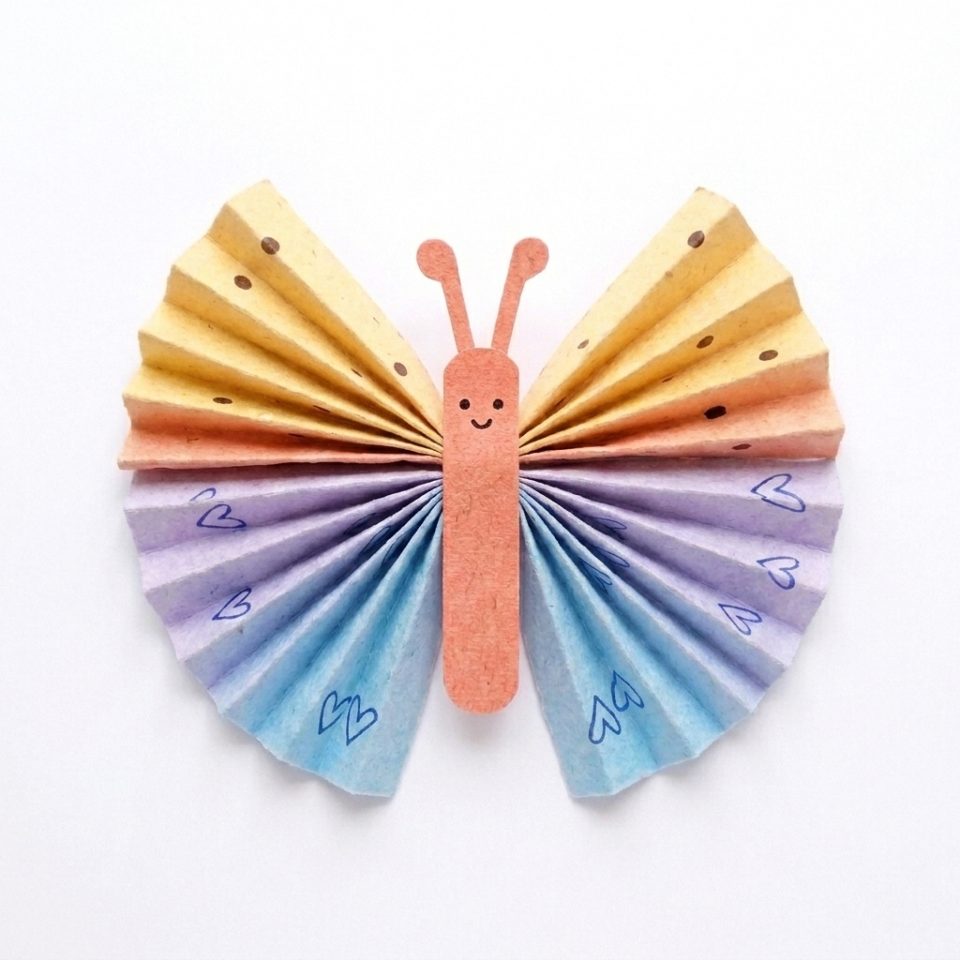 A finished paper butterfly with colorful fan wings, a coral body, a smile and rounded antennae.